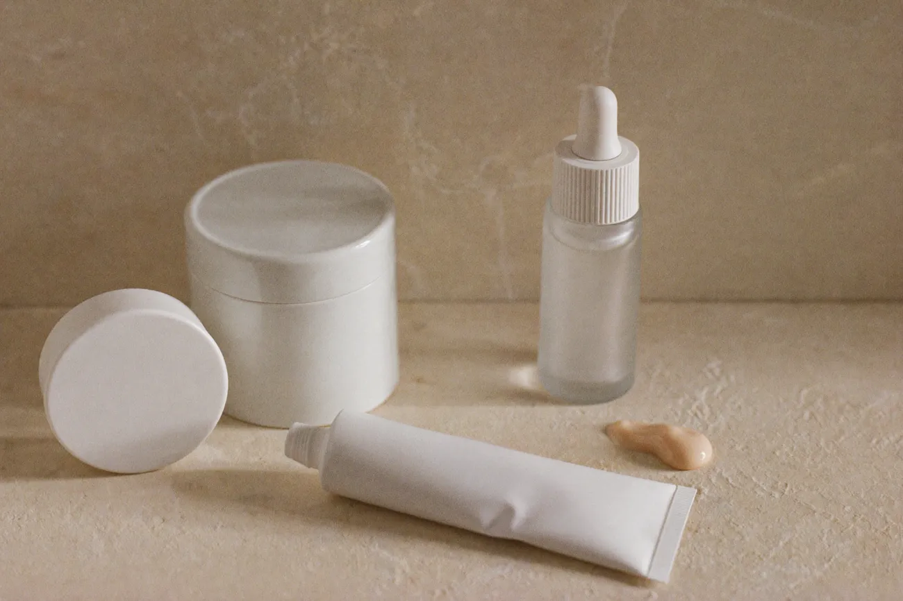 Drei unbeschriftete Pflegebehälter auf heller Steinfläche: Glasflasche mit Pipette, Keramiktiegel mit abgenommenem Deckel und liegende Tube.