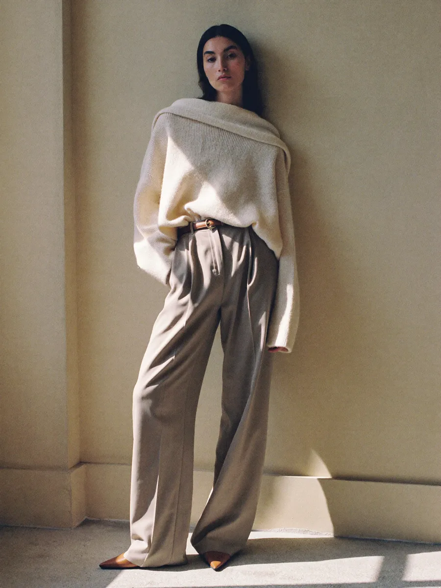 Frau in cremefarbenem Oversize-Kaschmirpullover und hoch geschnittener, weiter Hose vor einer hellen Wand.