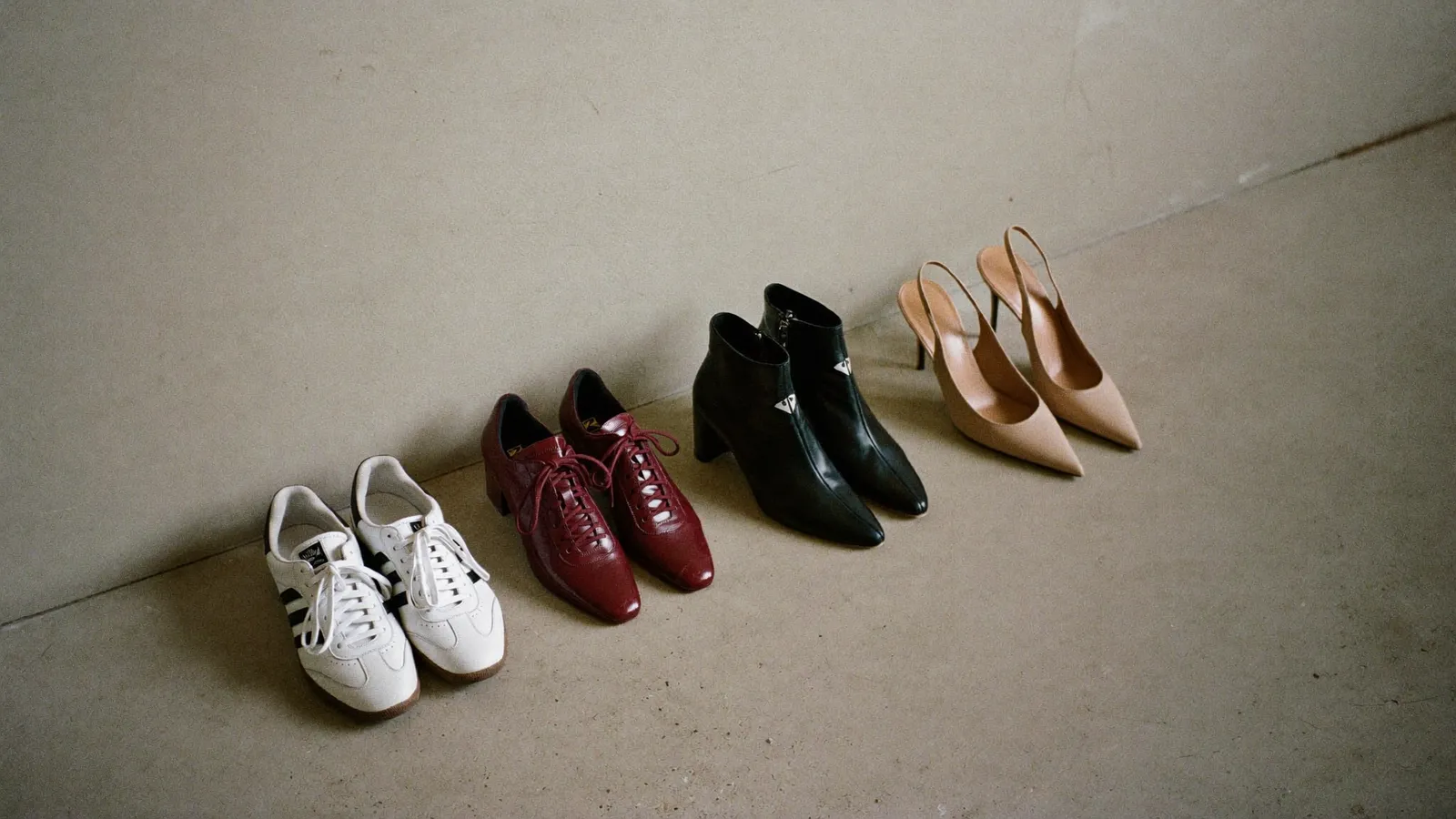 Vier Paar Damenschuhe nebeneinander auf hellem Betonboden: weiße Sneaker, burgunderrote Loafer, schwarze Stiefeletten und nudefarbene Slingbacks.