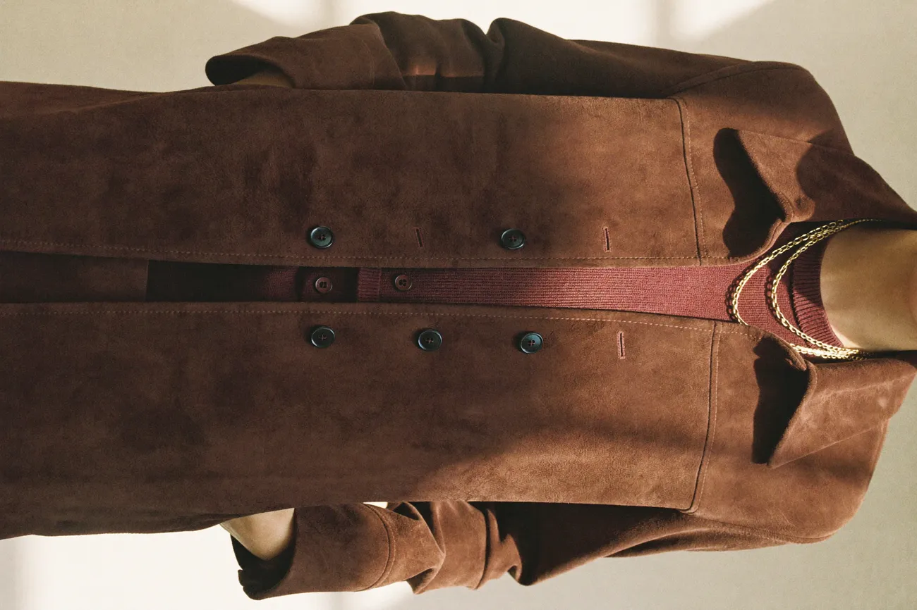 Nahaufnahme einer schokoladenbraunen Wildlederjacke über einem burgunderroten Feinstrickoberteil mit feinen Goldketten.